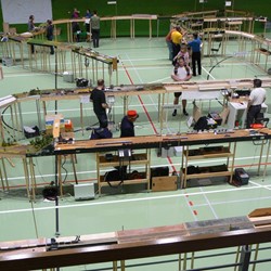 Pohled z galerie na posunující díl na svatoňovickém zhlaví  žst. Drnov, v druhém plánu na  žst. Výpravčice a s tratí do Červeného Kostelce s cukrovarem D. Cetno. Vlevo v pozadí Tomašov s odbočkou do Bocova, na obzoru pak trať z Tomašova přes Nový Oldřichov a Dírov do Mochova. Za povšimnutí stojí odborná debata nad technickým detailem v Dírově, čekání na křižování vlaků v Oldřichově, neboť vlak druhý právě vyjel z Tomašova a především s láskou, pečlivě a od útlého věku prováděná indoktrinace železničním modulářstvím u nevinného potomka strojvedoucím v Červeném Kostelci. Tahle fírová zřejmě po oválu jezdit nebude…